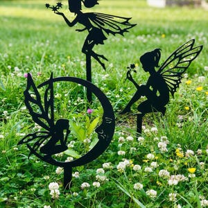 May include: Black metal fairy garden stakes. The set features three different fairy designs: one holding stars, one seated with a flower, and one within a crescent moon. These stakes are designed for placement in a garden or lawn.
