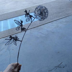 May include: A black metal sculpture depicting a dandelion with three fairy figures. The fairies are positioned on the stem and the dandelion head. The sculpture is held in a hand against a grey and blue background.