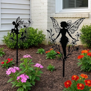 May include: Two black metal fairy garden stakes. One shows a fairy on a dandelion, the other a fairy holding flowers. The stakes are in a garden bed with pink and orange flowers.