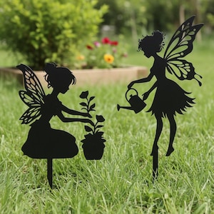 May include: Two black metal fairy garden stakes. One fairy kneels tending a potted plant, the other waters a plant with a watering can. Both fairies have detailed wings and are set in a grassy garden.