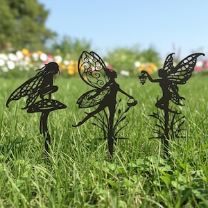 May include: Three black metal fairy garden stakes. One fairy sits on a mushroom, another holds a basket, and the third holds a lantern. The stakes are placed in a grassy area with flowers in the background.