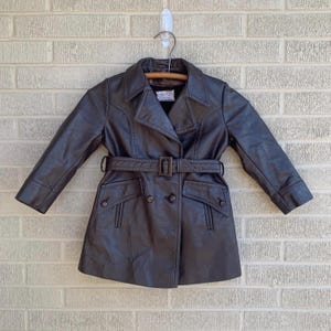 May include: A brown faux leather trench coat with a belt, double breasted closure, and two flap pockets.