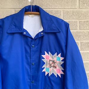 Può includere: Una giacca blu con un design a stella patchwork sul davanti. La giacca ha una fodera in pile bianca e una chiusura a scatto.