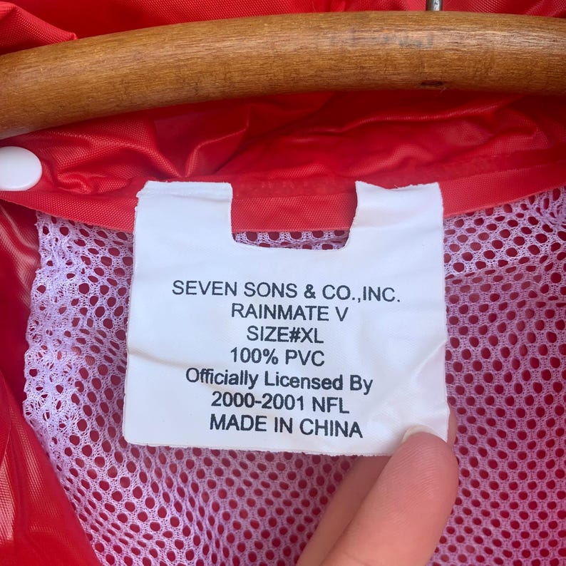 Puede incluir: Primer plano de un impermeable rojo con forro de malla blanca. Una etiqueta blanca muestra el texto "SEVEN SONS & CO., INC. RAINMATE V SIZE#XL 100% PVC Officially Licensed By 2000-2001 NFL MADE IN CHINA."
