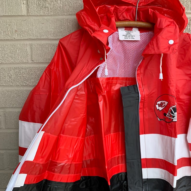 Puede incluir: Un impermeable de rayas rojas y blancas con capucha negra y cremallera negra. El impermeable tiene un forro de malla y un logotipo en blanco y negro de un casco de f&uacute;tbol con las letras "KC" en el lateral.