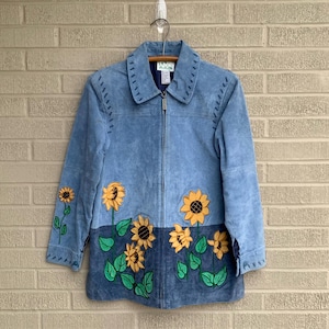 Könnte beinhalten: Eine blaue Wildlederjacke mit gestickten Sonnenblumen an den Ärmeln und vorne. Die Jacke hat einen Reißverschluss und einen Kragen.