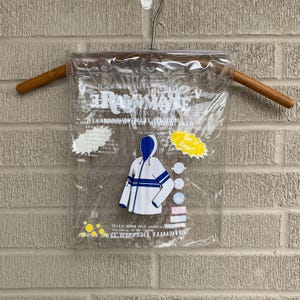 Puede incluir: Una bolsa de pl&aacute;stico transparente con cierre de cremallera, que contiene una chaqueta blanca con capucha y rayas azules. La chaqueta tiene el texto "Rainmate" impreso, junto con las palabras "Officially Licensed" y "Seven Sons and Company".