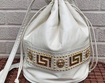 Puede incluir: Bolso de cuero color crema con cordón y adornos dorados. El bolso presenta una banda decorativa con tachuelas doradas, diseños de llaves griegas y medallones con cabeza de león. Cierre con cordón y correas para el hombro.