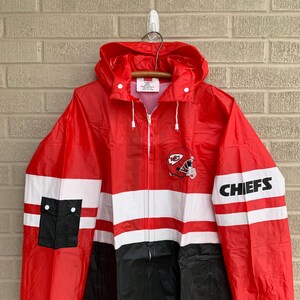 Puede incluir: Chaqueta de lluvia roja, blanca y negra con el logotipo de los Kansas City Chiefs y el texto "CHIEFS" en la manga. La chaqueta tiene capucha y cierre de cremallera.