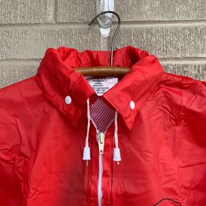 Puede incluir: Chaqueta roja de nylon con cremallera blanca y botones blancos. La etiqueta dice "Rainmate V Talla L 100% Nylon Oficialmente Licenciado Por 2007 NFL Hecho en China."