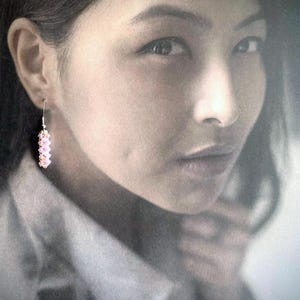 Peut inclure: Gros plan sur le visage d'une femme avec une boucle d'oreille en perles roses. La femme porte une chemise blanche et a les cheveux tirés en arrière.