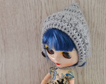 Op de afbeelding: Een pop met blauw haar die een grijze gehaakte hoed en een bloemenjurk draagt. De pop zit op een bruine doos met een houten ring die aan een touwtje is bevestigd.