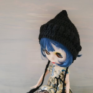 May include: A blue-haired doll wearing a black knitted hat with long ties, a floral dress, and grey lace-up boots. The doll is sitting on a white surface.
