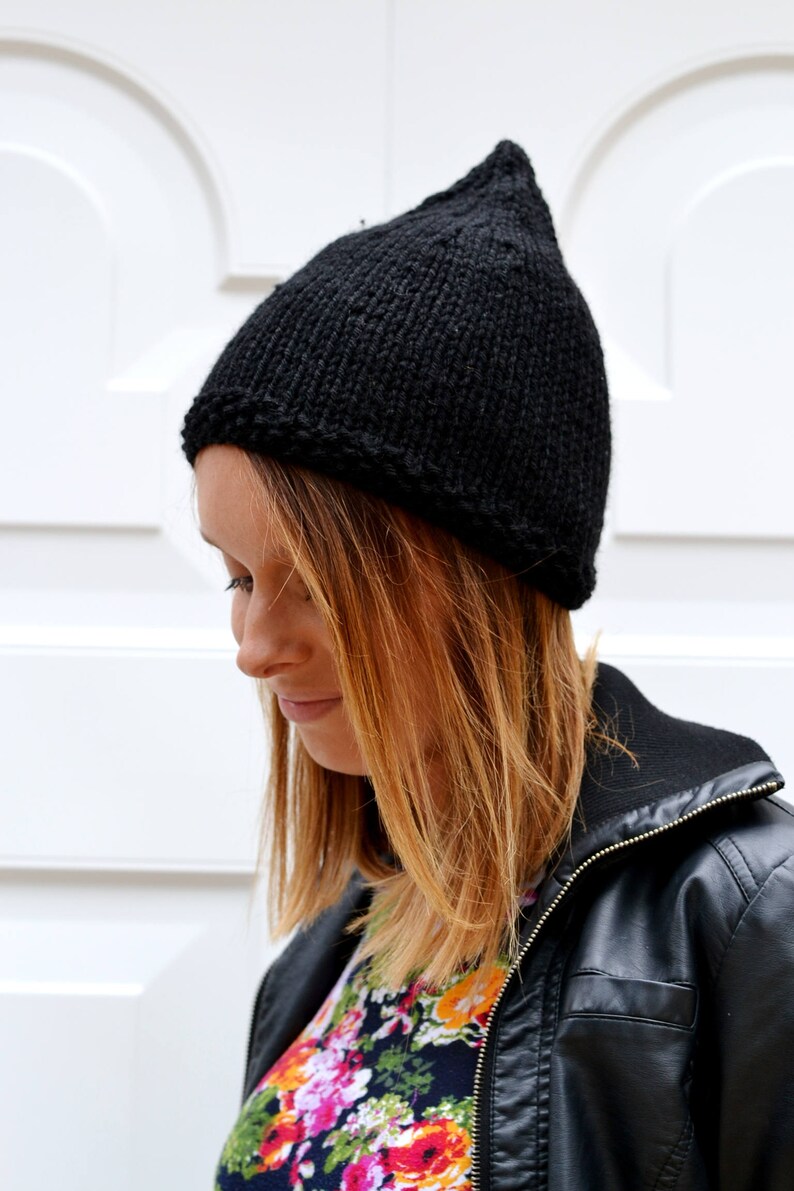 Op de afbeelding: Een zwarte gebreide beanie met een puntige top. De beanie wordt gedragen door een persoon met lang, lichtbruin haar. De persoon draagt een zwart leren jack en een shirt met bloemenprint.