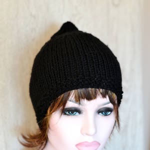 Op de afbeelding: Een zwarte gebreide beanie met een puntige top. De beanie wordt gedragen door een mannequin met bruin haar en blauwe ogen.