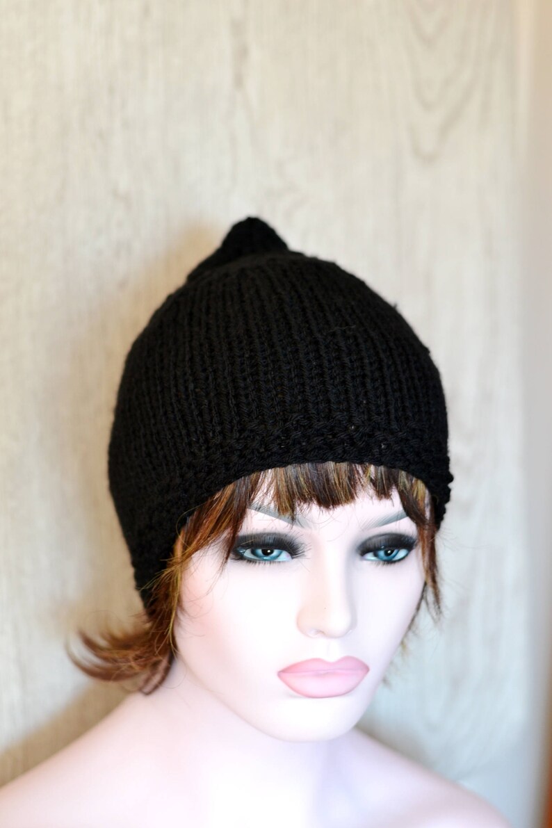Op de afbeelding: Een zwarte gebreide beanie met een puntige top. De beanie wordt gedragen door een mannequin met bruin haar en blauwe ogen.