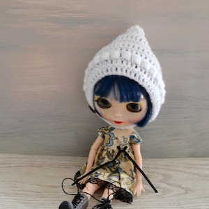 May include: A doll with blue hair wearing a white crocheted hat and a floral dress. The doll is sitting on a wooden surface.