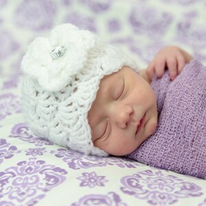 Op de afbeelding: Een slapende baby die een witte gehaakte muts met een bloemdetail en een paarse swaddle draagt. De baby ligt op een wit en paars gestreept deken.