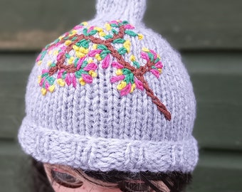 Op de afbeelding: Een grijze gebreide muts met een bruin geborduurde tak met roze en gele bloemen. De muts zit op een pop met zwart haar en een witte jurk.