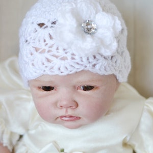 Op de afbeelding: Een witte gehaakte baby muts met een witte bloem en een strassversiering.