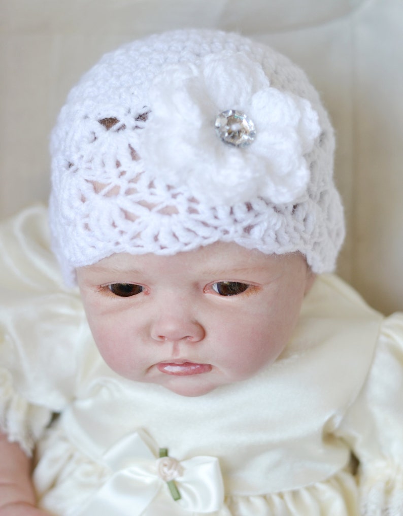 Op de afbeelding: Een witte gehaakte baby muts met een witte bloem en een strassversiering.