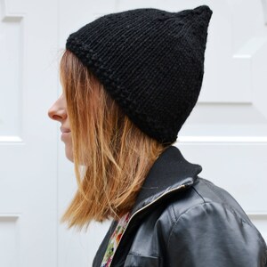 Op de afbeelding: Een zwarte gebreide beanie met een puntige top. De beanie wordt gedragen door een persoon met bruin haar en een zwart leren jack.