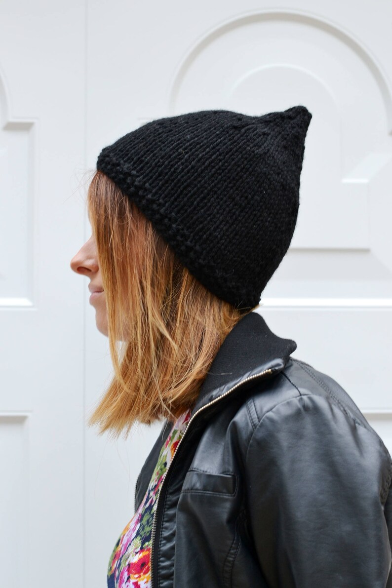 Op de afbeelding: Een zwarte gebreide beanie met een puntige top. De beanie wordt gedragen door een persoon met bruin haar en een zwart leren jack.