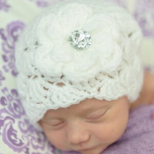Op de afbeelding: Een witte gehaakte baby muts met een grote witte bloem en een glinsterende strass versiering. De muts zit op het hoofd van een slapende baby.