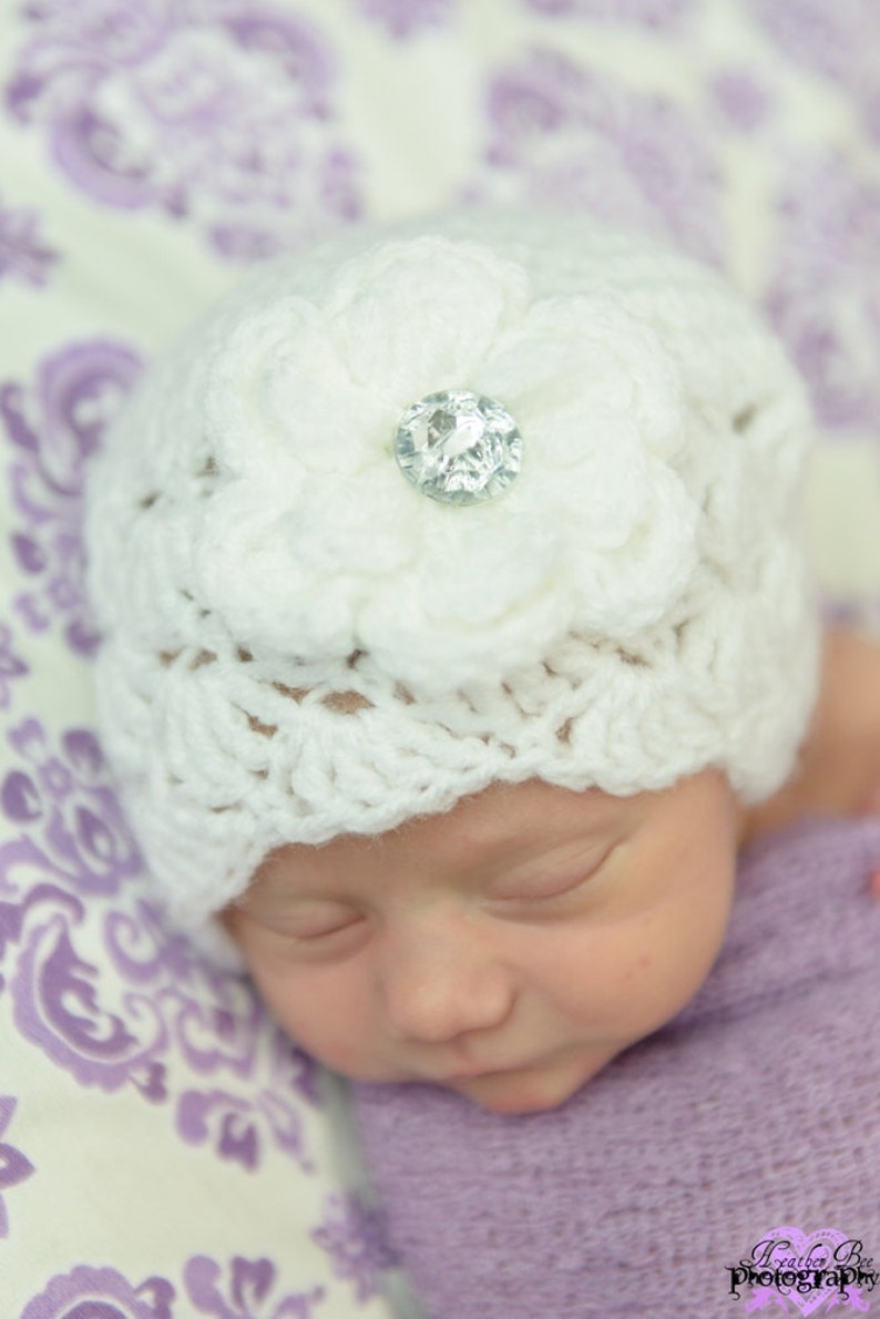Op de afbeelding: Een witte gehaakte baby muts met een grote witte bloem en een glinsterende strass versiering. De muts zit op het hoofd van een slapende baby.