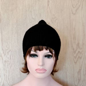 Op de afbeelding: Een zwarte gebreide beanie met een simpel ontwerp. De muts wordt gedragen door een mannequin met bruin haar en een neutrale uitdrukking.