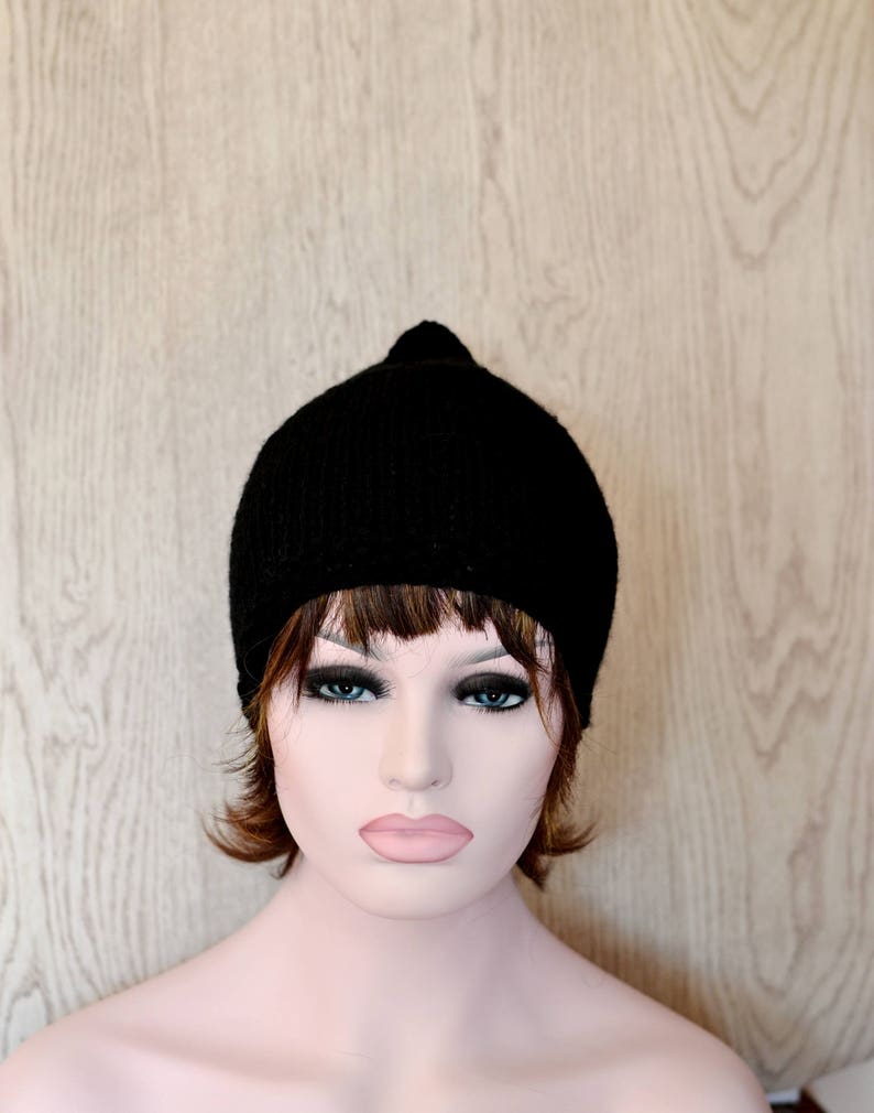 Op de afbeelding: Een zwarte gebreide beanie met een simpel ontwerp. De muts wordt gedragen door een mannequin met bruin haar en een neutrale uitdrukking.
