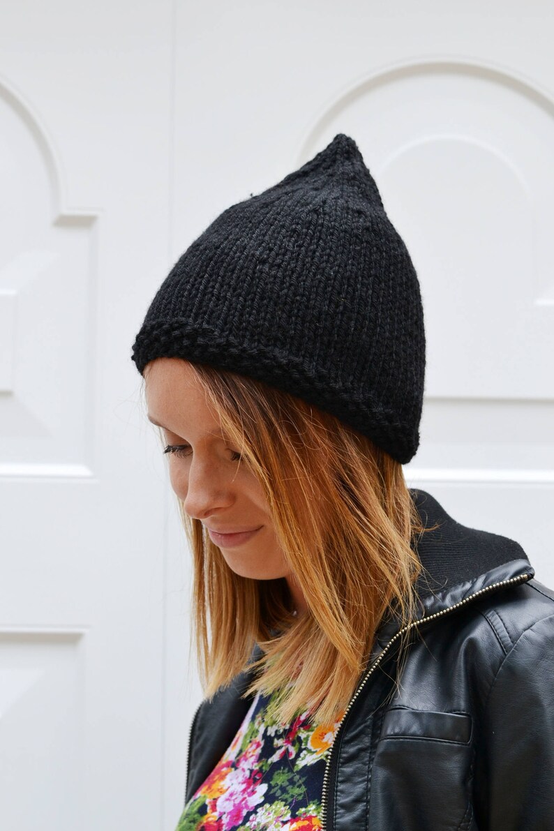 Op de afbeelding: Een zwarte gebreide beanie met een puntige top. De beanie wordt gedragen door een persoon met lang, bruin haar.