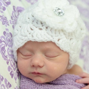 Op de afbeelding: Een slapende baby die een witte gehaakte muts draagt met een bloem en een strassversiering. De muts is op een paarse en witte gepatroneerde achtergrond.