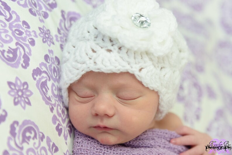 Op de afbeelding: Een slapende baby die een witte gehaakte muts draagt met een bloem en een strassversiering. De muts is op een paarse en witte gepatroneerde achtergrond.
