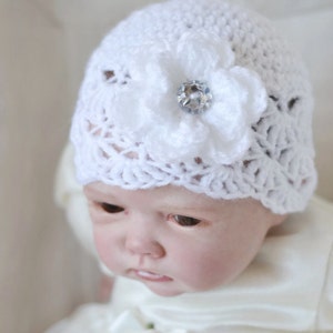 Op de afbeelding: Een witte gehaakte baby muts met een witte gehaakte bloem en een heldere strass steentje in het midden.