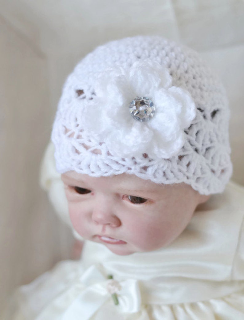 Op de afbeelding: Een witte gehaakte baby muts met een witte gehaakte bloem en een heldere strass steentje in het midden.