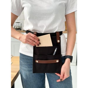 Puede incluir: Un delantal de lona negro con correas de cuero marrón y un asa de cuero marrón. El delantal tiene varios bolsillos, incluido un bolsillo grande con un cuaderno en su interior y un bolsillo más pequeño con un bolígrafo.