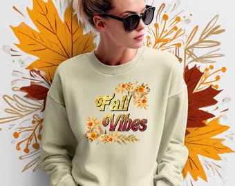 May include: A cream-colored sweatshirt with the text "Fall Vibes" in a decorative font, accented with floral and leaf designs. The background features autumn leaves in shades of orange and brown.