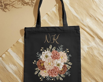 Könnte beinhalten: Schwarze Stofftasche mit einem Blumendesign in Rosa-, Weiß- und Brauntönen. Die Tasche hat ein goldenes Monogramm "NK" auf der Vorderseite.