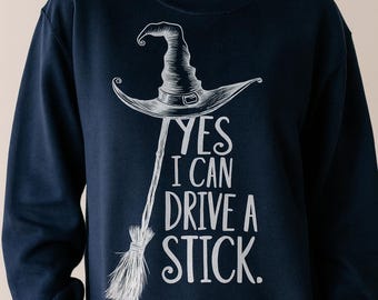 May include: Navy blue sweatshirt with a white graphic of a witch's hat and broom. The text "YES I CAN DRIVE A STICK" is printed below the hat. A casual, comfortable top for Halloween or everyday wear.