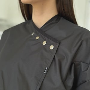 Può includere: Un mantello da salone nero con un design avvolgente e chiusure a scatto argentate. Il mantello ha un colletto alto e una piccola etichetta con la scritta "PROF KIT". Il tessuto sembra essere un materiale liscio e opaco.