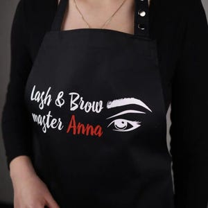 May include: Black apron with white text that reads "Lash & Brow master Anna" and a white stylized eye with an eyebrow.
