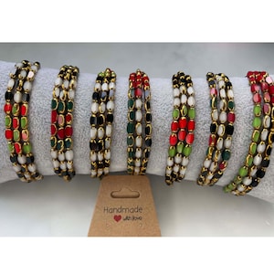Könnte beinhalten: Sechs bunte Perlenarmbänder mit einem Mix aus roten, grünen, schwarzen und weißen Perlen. Die Armbänder sind auf einer weißen Oberfläche gestapelt und haben einen goldfarbenen Metalldraht, der durch sie verläuft. Der Text "Handmade with Love" ist auf einem braunen Etikett gedruckt.