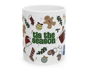 May include: White ceramic mug decorated with Christmas-themed illustrations, including a gingerbread man, stocking, and a Santa hat. The mug features the text "tis the season" in green letters, perfect for holiday beverages.