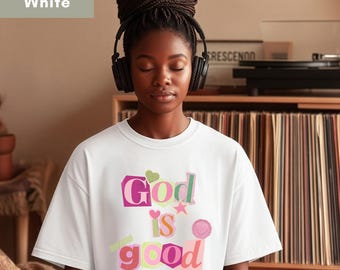 May include: A white short-sleeved t-shirt featuring a multi-coloured graphic print with the text God is good. The lettering uses various fonts and colours including pink, orange, and green, accented by small shapes like a heart, a star, and a circle.