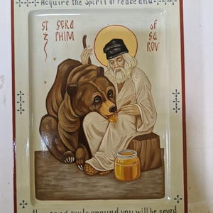 Może przedstawiać: Oprawiona ikona religijna przedstawiająca świętego i niedźwiedzia. Święty, w czarnym kapeluszu i białych szatach, dzieli się miodem z niedźwiedziem. Rama zawiera tekst: "Zdobądź Ducha Pokoju i... tysiąc dusz wokół ciebie zostanie zbawionych."