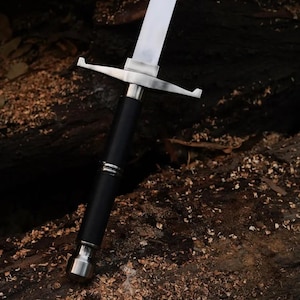 Puede incluir: Una daga de plata con mango negro y guarda cruzada. La daga está apoyada sobre una superficie de madera marrón oscura.