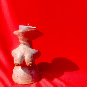 Puede incluir: Un portavelas de cerámica con forma de torso de mujer con una vela de té encendida en la parte superior. El portavelas es de color marrón claro con un acabado moteado y está sobre una superficie de tela roja.