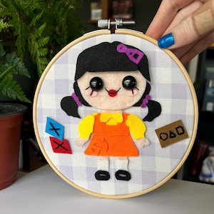 May include: A felt doll with black hair, a purple bow, and a yellow and orange dress. The doll is holding a red and blue diamond shape and is standing on a white and purple gingham background. The doll is framed in a wooden hoop.