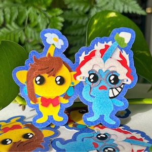 May include: A collection of colorful felt character stickers. One is yellow with brown hair and a red bow tie. Another is blue with white and red hair, wearing glasses. The stickers have a blue outline and are set against a green leafy background.
